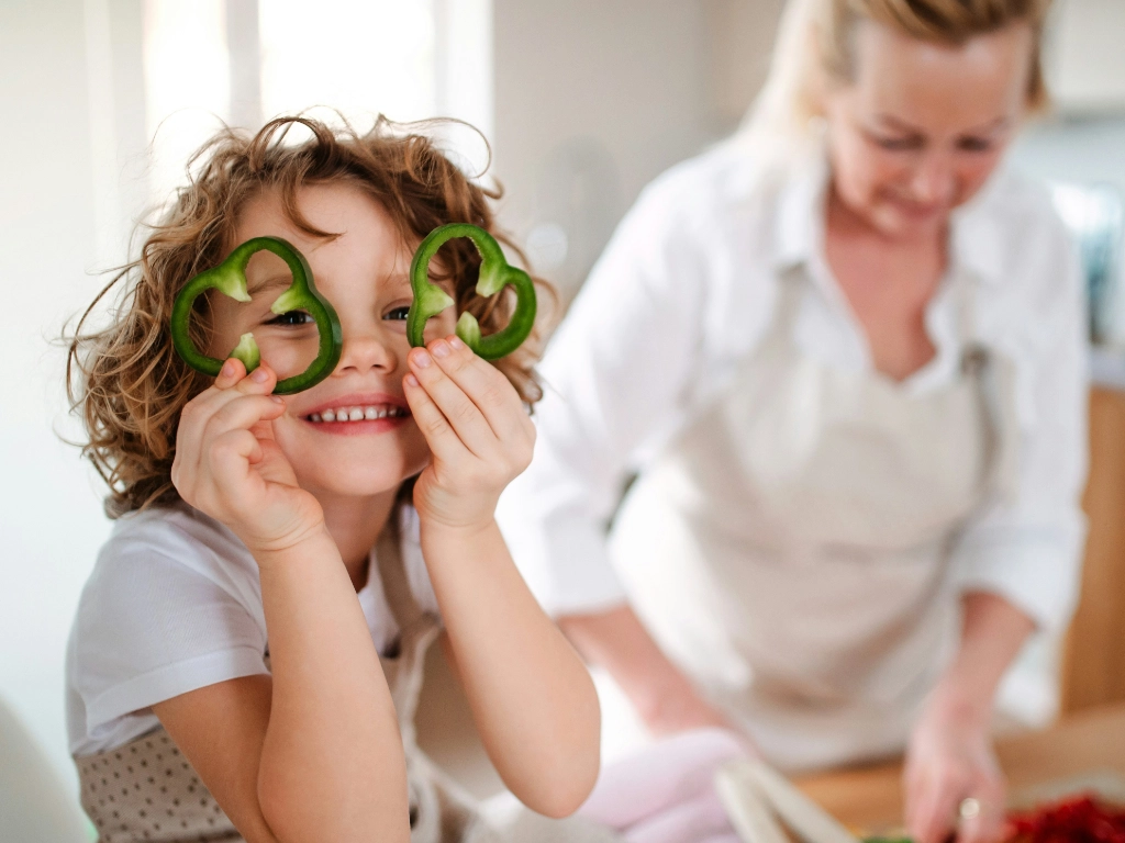 Mutter schnipselt Gemüse und das Kind legt Paprika-Ringe um die Augen die aussehen wie Herzen.